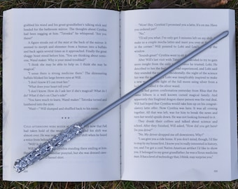 May include: A silver wand with a sparkly, textured surface. The wand is lying on an open book with text about a magical creature. The book is on a bed of green grass.