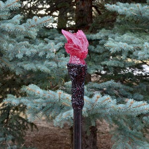 May include: A decorative staff with a dark, textured handle and a vibrant pink, flame-shaped top. The staff is set against a backdrop of blue-green evergreen trees, creating a striking contrast.