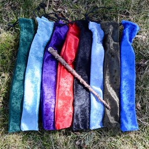 May include: Eight velvet pouches in various colors, including green, blue, purple, red, black, and dark blue, are laid out on a bed of green grass. A wooden wand with a brown leather wrap is lying on top of the pouches.