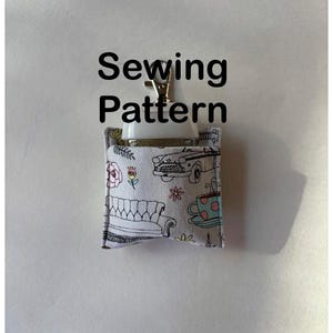 Pode incluir: Um pequeno porta-padrões de costura de tecido com um fecho de metal. O suporte é branco com um padrão de sofá, carro, xícara de chá e flores. As palavras "Sewing Pattern" são impressas em preto.