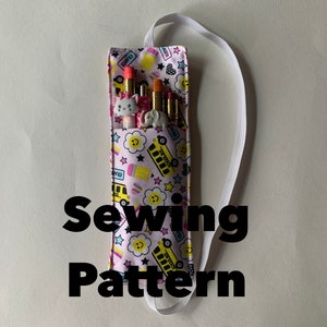 以下が含まれることがあります： 白いストラップが付いたピンクの鉛筆ケース。ケースにはスクールバスとスマイリーフェイスのプリントが施されています。ケースには「SCHOOL BUS」の文字が印刷されています。