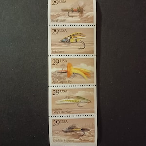 Può includere: Cinque francobolli americani vintage con illustrazioni di diverse mosche da pesca. Ogni francobollo ha il testo "29 USA" e il nome della mosca, tra cui "Royal Wulff", "Jock Scott", "Apte Tarpon Fly", "Lefty's Deceiver" e "Muddler Minnow".