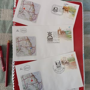 Può includere: Tre buste bianche con illustrazioni di mappe e francobolli, appoggiate su un quaderno rosso. Le buste presentano francobolli con testo e immagini. Una matita rossa e una rosa sono visibili.