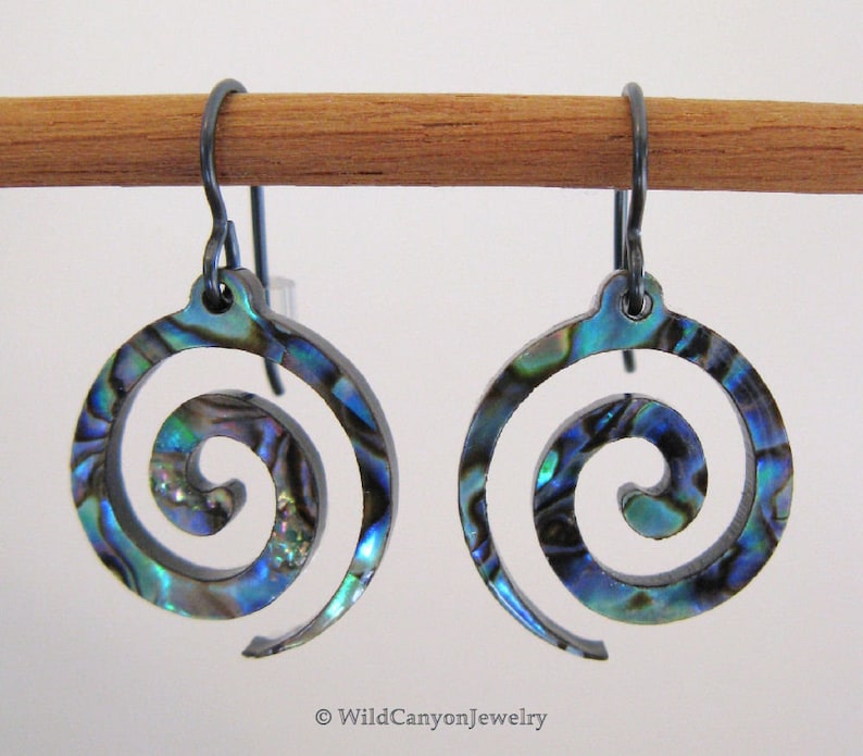 May include: A pair of earrings with a spiral design made from abalone shell. The earrings are a blue, green, and purple iridescent color. The earrings are hanging from black metal hooks.