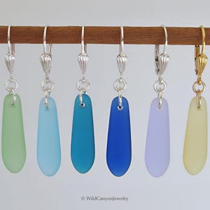 May include: A collection of six dangle earrings in various colors, including green, blue, and purple. Each earring features a smooth, elongated glass pendant suspended from a silver or gold-colored hook. The earrings are displayed on a wooden rod.