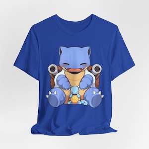 May include: A royal blue short-sleeve t-shirt featuring a graphic print of a large light blue turtle character with two cannons on its back, sitting and holding a smaller light blue turtle character with a tan shell on its lap.