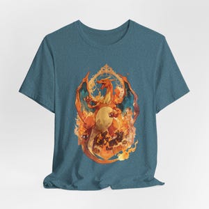 May include: A heathered teal short-sleeve t-shirt featuring a central graphic of a large orange dragon-like creature with wings spread, surrounded by stylized flames and enclosed within an ornate, decorative golden-colored frame design.