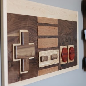 May include: A wooden wall art piece in the shape of a classic video game controller. The controller is made of different types of wood, including walnut and maple. The buttons are made of red wood.