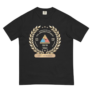 Puede incluir: Camiseta negra con un emblema dorado. El emblema incluye las palabras "UNITED STATES ARMY", "2ND ARMORED DIVISION", "EST: 1940", "HELL ON WHEELS" y "COLD WAR VETERAN". La frase "HELL ON WHEELS" también se muestra.