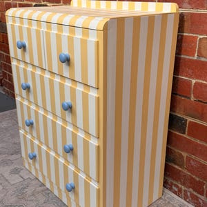 May include: A wooden chest of drawers featuring a vertical yellow and white striped pattern across the entire exterior. The piece includes five drawers, each equipped with two round, light blue knobs, and a flat top surface with rounded edges.