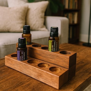 May include: A wooden essential oil holder with multiple circular openings, holding three amber glass bottles with black caps. The holder is made of light brown wood and sits on a wooden table. The bottles have labels with text.