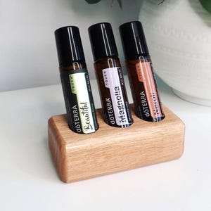 Op de afbeelding: Drie essentiële olie roll-on flessen in een houten houder. De flessen zijn gelabeld met "Beautiful", "Magnolia" en "Neroli". Elke fles heeft een zwarte dop en een etiket met de merknaam "dōTERRA". De houder is gemaakt van lichtgekleurd hout.