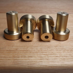 May include: Four brass cabinet knobs with a cylindrical body and a circular base. The knobs have a polished finish and are arranged on a wooden surface. The knobs are likely used for drawers or doors.