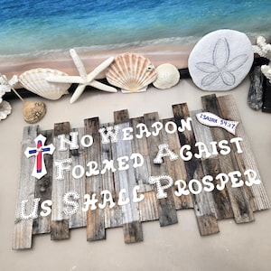 Könnte beinhalten: Ein rechteckiges Wandschild aus vertikalen, verwitterten Holzlatten mit dem weißen Text No Weapon Formed Against Us Shall Prosper. Ein Kreuz mit roter Mitte und ein kleines fischförmiges Schild mit dem Text Isaiah 54:17 sind daran befestigt.