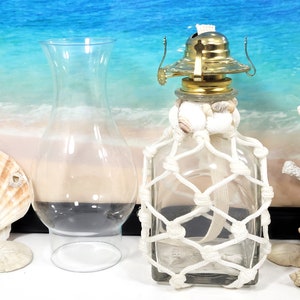 May include: A clear glass oil lamp with a white rope net and seashells around the base. The lamp is sitting on a white surface.