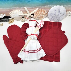 May include: A white kitchen towel with red stripes is folded into the shape of an angel. A wooden spoon with the text "God Bless Our Home" is attached to the angel. The angel is surrounded by red kitchen accessories, including a pot holder and oven mitt.