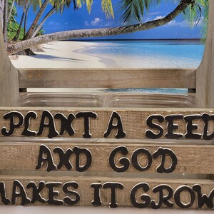 May include: Wooden planter box with the text "I PLANT A SEED AND GOD MAKES IT GROW" in black letters. The box has a handle and two clear plastic containers inside. The background shows a beach scene.