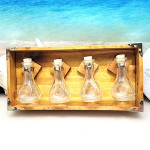 May include: A wooden box with four clear glass bottles with cork stoppers. The bottles are mounted on wooden supports inside the box. The box has metal corner reinforcements. The background features a beach scene with blue water.
