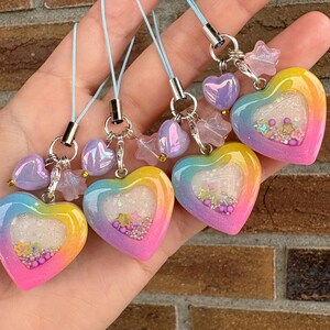 May include: Four heart-shaped charms with a rainbow gradient design, filled with glitter and small embellishments. Each charm is attached to a light blue cord with a silver-coloured clasp and decorative beads. The charms are held in a hand.