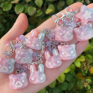 May include: A selection of pastel resin charms in the shapes of bears, bunnies, cats, and a star. They are attached to silver chains with lobster clasps and embellished with pink and purple beads. The charms have a glittery, iridescent finish.