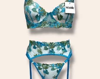 May include: A matching lingerie set featuring a blue underwire bra and a high-waisted garter belt with a thong. Both pieces are crafted from sheer white mesh fabric adorned with intricate blue and green floral embroidery. A tag reads Scantilly.