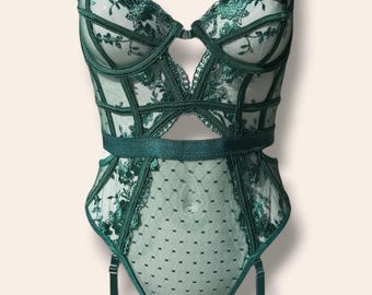 May include: A dark emerald green bodysuit featuring sheer mesh panels with intricate floral embroidery and a structured cage-style bodice. The garment includes adjustable straps, a cutout waist, and attached garter straps. A white tag reads scantie.