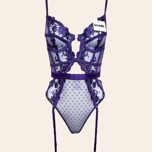 May include: A purple lingerie set featuring a sheer bodice with floral embroidery and a cut-out design. The set includes a matching garter belt and thong, with adjustable straps. The tag reads "Scantee".