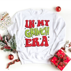 May include: A white sweatshirt with the text "IN MY GRINCH ERA" in red and green. The sweatshirt is surrounded by Christmas decorations, including ornaments, gift boxes, and greenery.