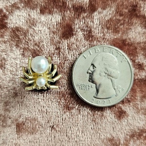 May include: A gold-colored spider brooch with a pearl body and a quarter coin for size comparison. The spider has eight legs and a black base. The brooch is on a pinkish-brown velvet surface.