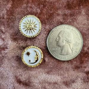 May include: Two celestial-themed buttons with gold and white enamel designs. One button features a sun, the other a crescent moon and star. A U.S. quarter is next to the buttons for size comparison.