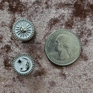 以下が含まれることがあります： 銀色の縁取りが付いた白いエナメル製のピンが3つ。ピンには太陽、星が1つの三日月、星が2つの三日月が描かれている。ピンは茶色の布地に置かれている。