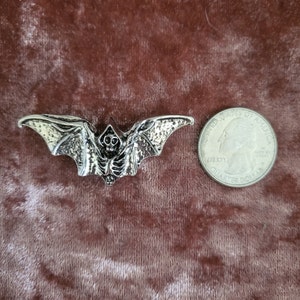 May include: Silver-toned metal bat brooch with a skull and ribcage design. The brooch is about the size of a US quarter.