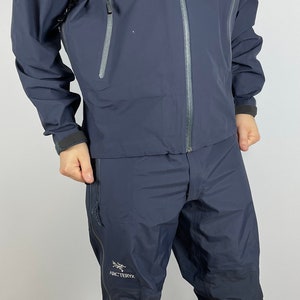 Könnte beinhalten: Ein dunkelblauer wasserdichter Jacke- und Hosen-Set von Arc'teryx. Die Jacke hat eine Kapuze und einen Reißverschluss. Die Hose hat eine Reißverschlusstasche am linken Bein.