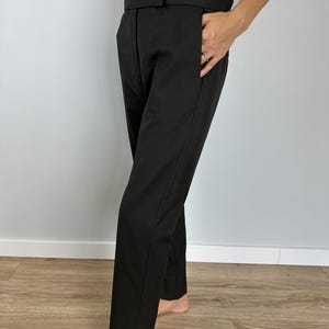 Puede incluir: Pantalones negros a medida con pernera recta. Los pantalones tienen cintura alta y bolsillos. La tela parece ser de color liso y uniforme. Los pantalones se llevan con una camiseta blanca.