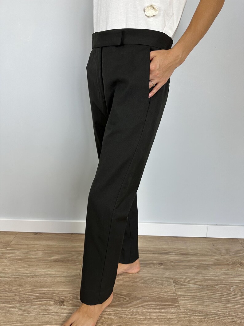 Puede incluir: Pantalones negros a medida con pernera recta. Los pantalones tienen cintura alta y bolsillos. La tela parece ser de color liso y uniforme. Los pantalones se llevan con una camiseta blanca.
