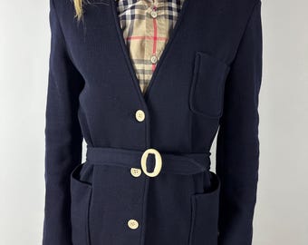 Peut inclure: Un cardigan bleu marine avec une ceinture assortie et une boucle crème. Le cardigan a deux poches avant et des boutons crème. Une chemise à carreaux est visible en dessous.