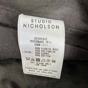 Puede incluir: Primer plano de una etiqueta blanca con texto negro de Studio Nicholson. La etiqueta detalla la composici&oacute;n de la tela: 60% lana virgen, 40% poliamida y forro de 50% cupro y 50% algod&oacute;n. Incluye instrucciones de cuidado.