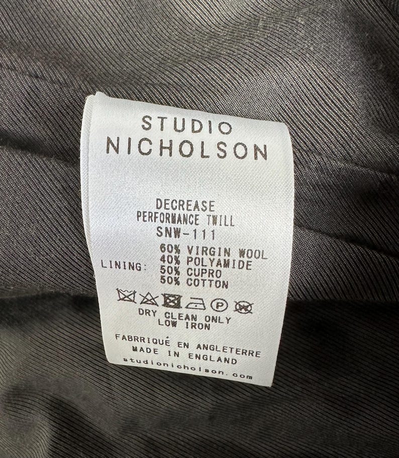 Puede incluir: Primer plano de una etiqueta blanca con texto negro de Studio Nicholson. La etiqueta detalla la composici&oacute;n de la tela: 60% lana virgen, 40% poliamida y forro de 50% cupro y 50% algod&oacute;n. Incluye instrucciones de cuidado.