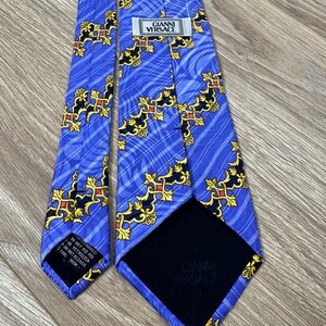 以下が含まれることがあります： 金色と黒の模様が施された青いシルクネクタイ。ネクタイには「GIANNI VERSACE」と書かれたラベルが付いています。