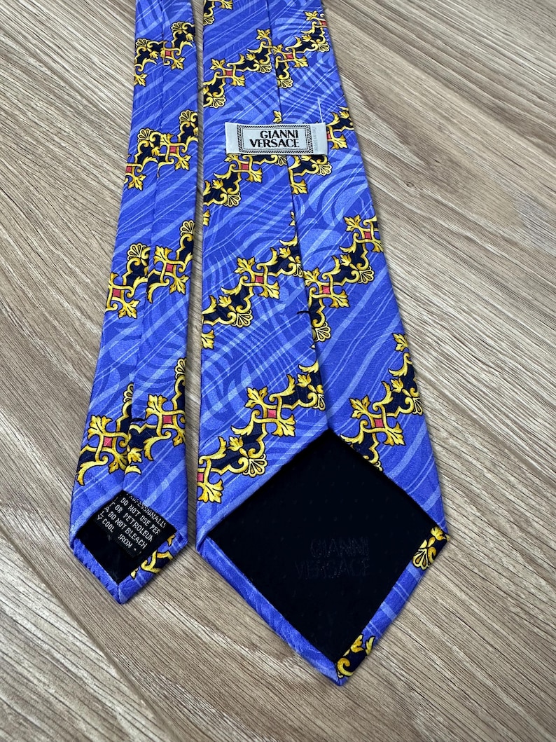 以下が含まれることがあります： 金色と黒の模様が施された青いシルクネクタイ。ネクタイには「GIANNI VERSACE」と書かれたラベルが付いています。