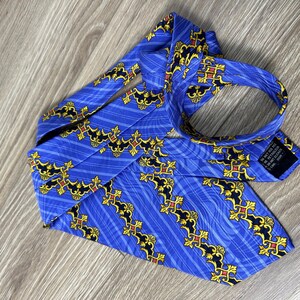 以下が含まれることがあります： 金色と黒の模様が施された青いシルクネクタイ。模様は、スタイリッシュな花の形と線が繰り返されるモチーフになっています。ネクタイは幅が狭く、先端が尖っています。