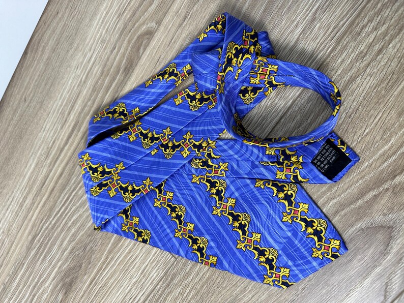 以下が含まれることがあります： 金色と黒の模様が施された青いシルクネクタイ。模様は、スタイリッシュな花の形と線が繰り返されるモチーフになっています。ネクタイは幅が狭く、先端が尖っています。