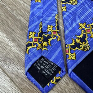 以下が含まれることがあります： 金色とピンクの模様が施された青いシルクネクタイ。ネクタイには、"Professionally Dry Clean Only, Do Not Use Per Or Petroleum, Do Not Bleach, Cool Iron."というテキストが書かれた黒いラベルが付いています。