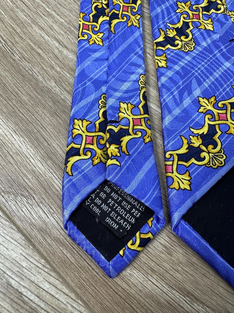 以下が含まれることがあります： 金色とピンクの模様が施された青いシルクネクタイ。ネクタイには、"Professionally Dry Clean Only, Do Not Use Per Or Petroleum, Do Not Bleach, Cool Iron."というテキストが書かれた黒いラベルが付いています。