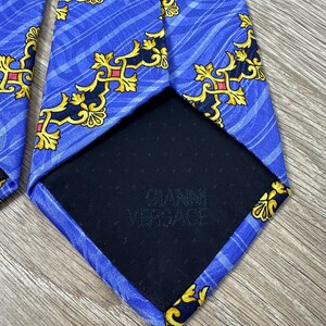 以下が含まれることがあります： 金色と黒の模様が施された青いシルクネクタイ。ネクタイの裏には「Gianni Versace」の文字が印刷されています。