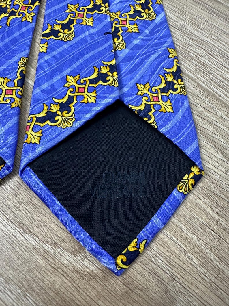 以下が含まれることがあります： 金色と黒の模様が施された青いシルクネクタイ。ネクタイの裏には「Gianni Versace」の文字が印刷されています。