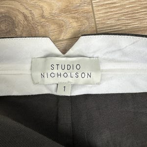 Puede incluir: Primer plano del interior de la cintura de un pantal&oacute;n gris oscuro. La etiqueta blanca dice "STUDIO NICHOLSON" con el n&uacute;mero "1" debajo. El pantal&oacute;n est&aacute; hecho de una tela texturizada.