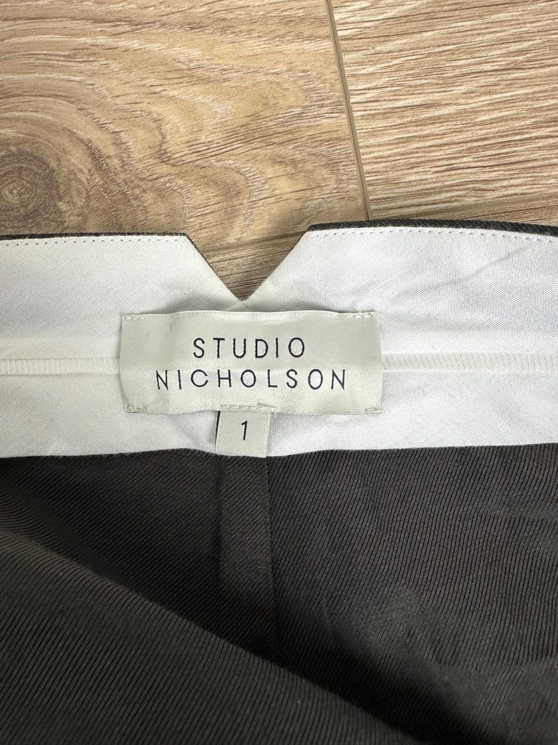 Puede incluir: Primer plano del interior de la cintura de un pantal&oacute;n gris oscuro. La etiqueta blanca dice "STUDIO NICHOLSON" con el n&uacute;mero "1" debajo. El pantal&oacute;n est&aacute; hecho de una tela texturizada.