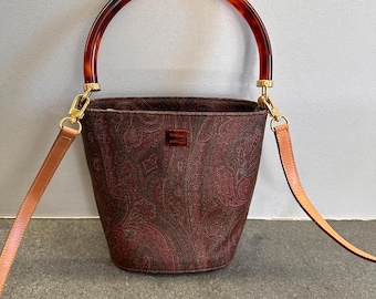 May include: A bucket bag featuring a dark red and black paisley pattern, a rounded tortoiseshell-style handle, and a detachable tan leather shoulder strap. The bag includes gold-tone metal hardware and a small rectangular logo plaque on the front.