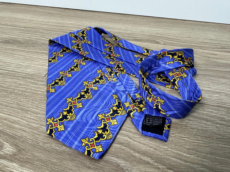 以下が含まれることがあります： 金色と黒色の模様が施された青いシルクネクタイ。ネクタイには、青と黒のラベルが付いており、「ドライクリーニング不可、石油系ドライクリーニングのみ、イタリア製」と記載されています。